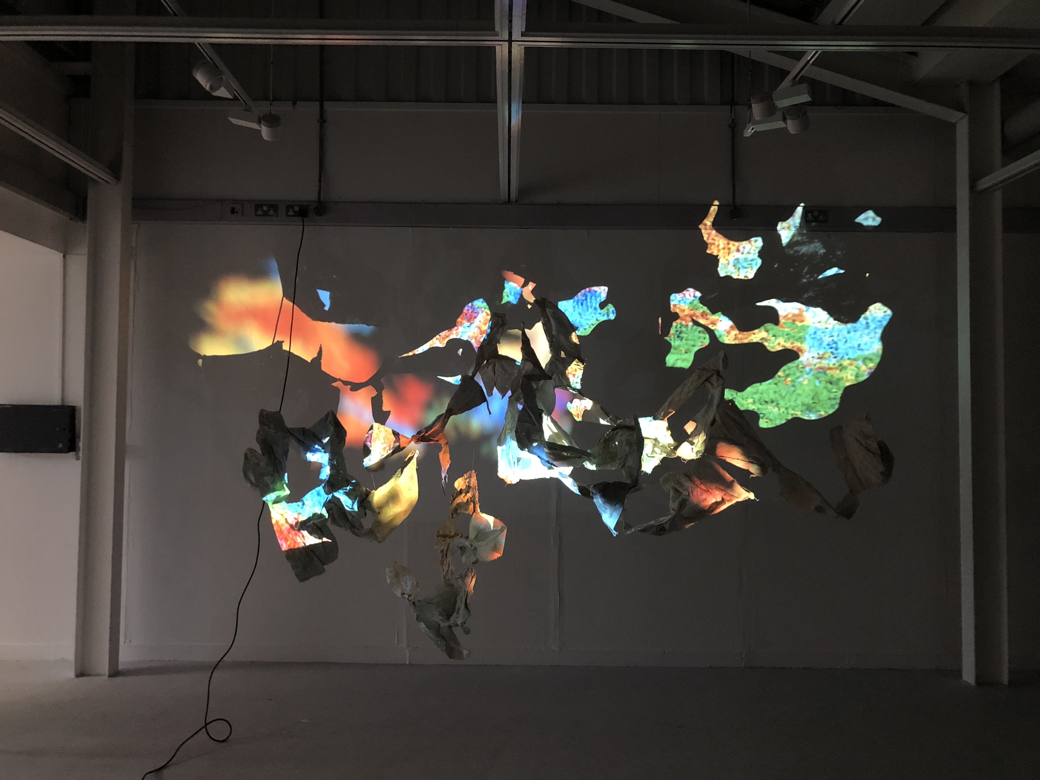 An image of a cluster of hanging forms, which weave in and out of each other, are being projected onto with distorted, vibrant images which "paint" the surface of the hanging sculptures