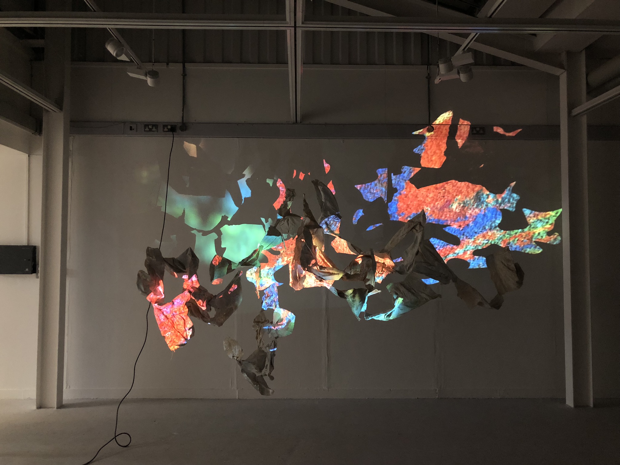 An image of a cluster of hanging forms, which weave in and out of each other, are being projected onto with distorted, vibrant images which "paint" the surface of the hanging sculptures
