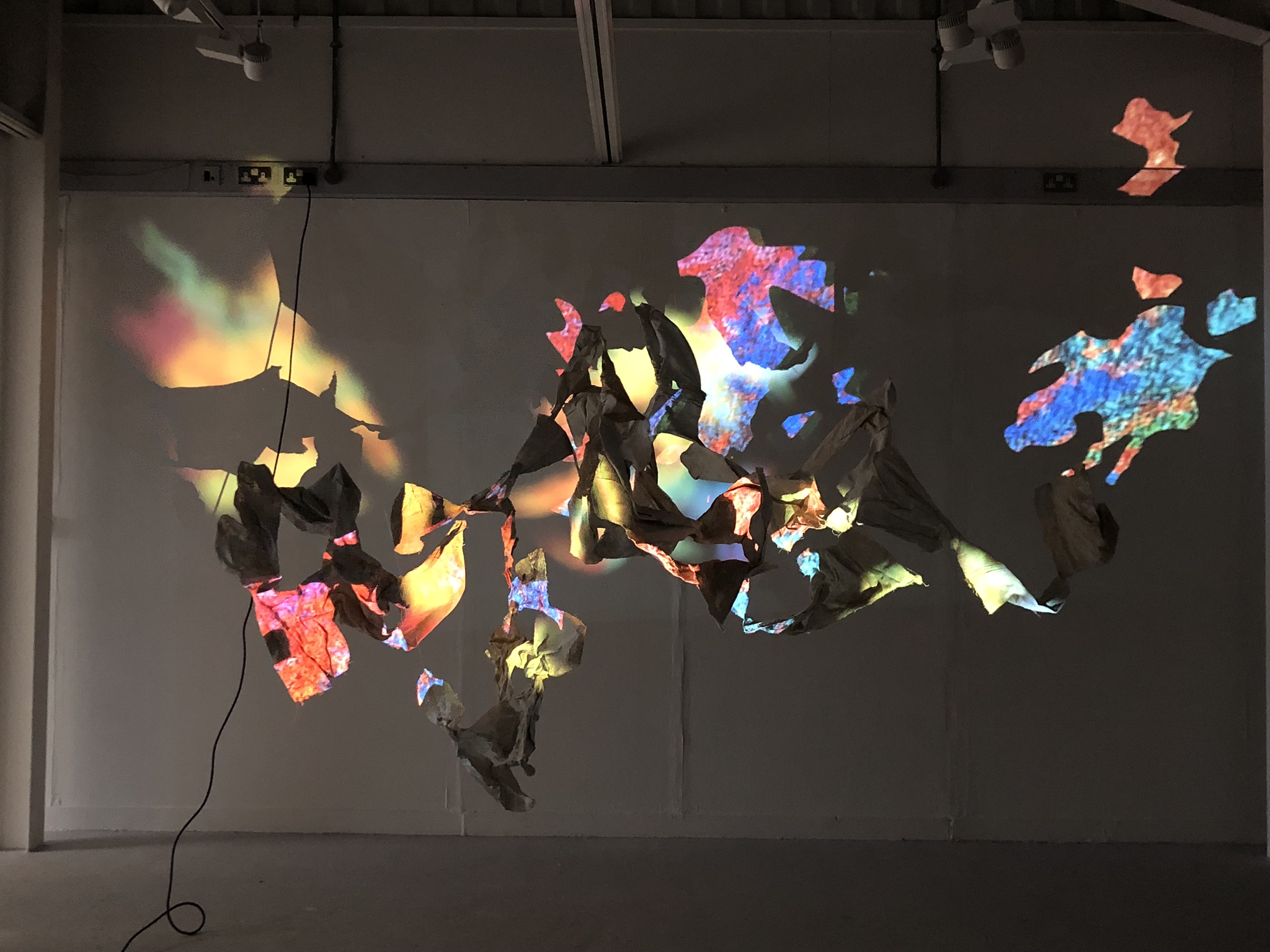 An image of a cluster of hanging forms, which weave in and out of each other, are being projected onto with distorted, vibrant images which "paint" the surface of the hanging sculptures