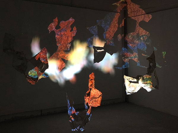 An image of colourful, distorted projected images which are being projected onto fabric forms which hang in the centre of the space