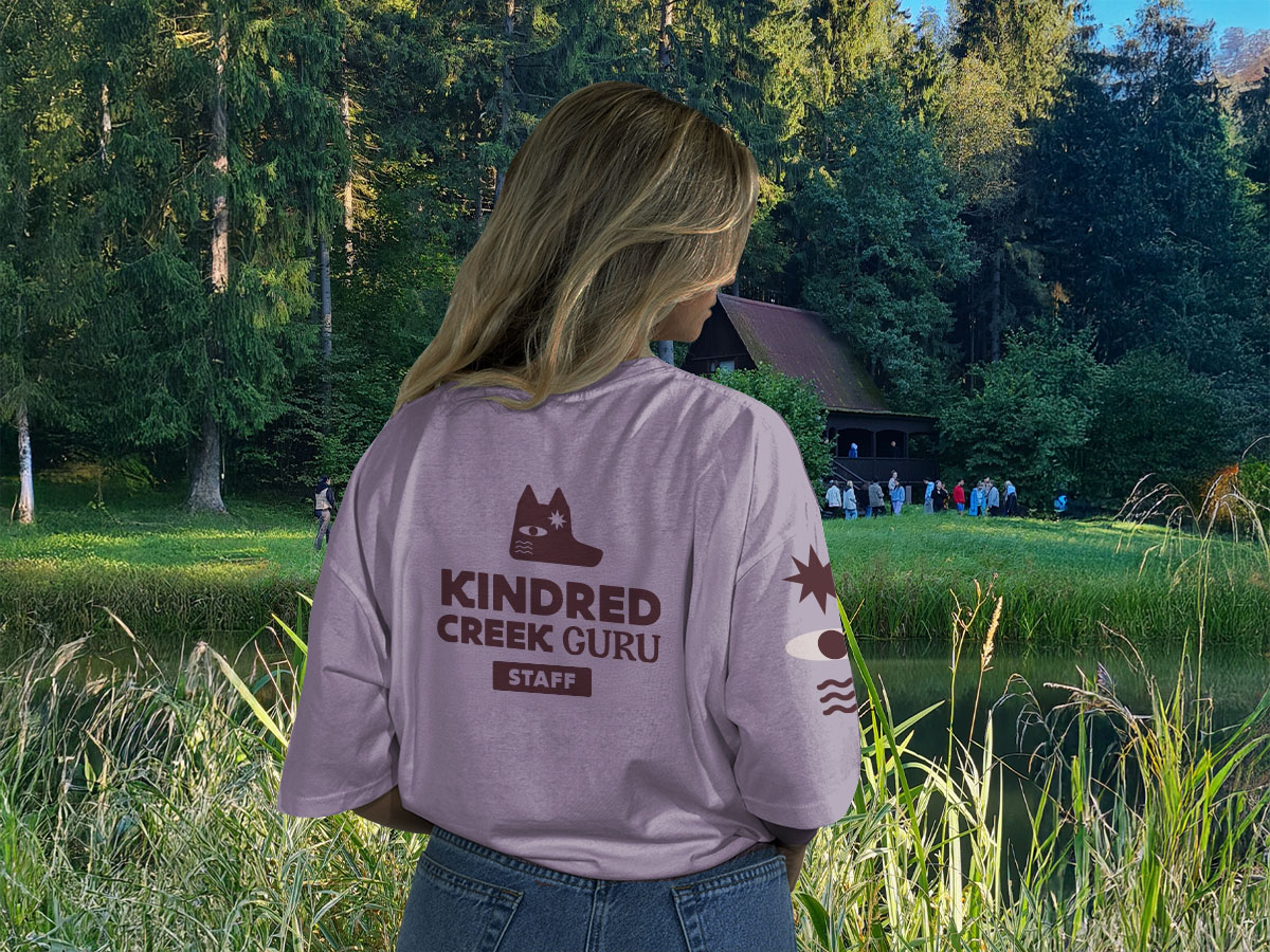 A photograph of a woman from behind looking across a lake while wearing a purple t-shirt that says Kindred Creek guru.