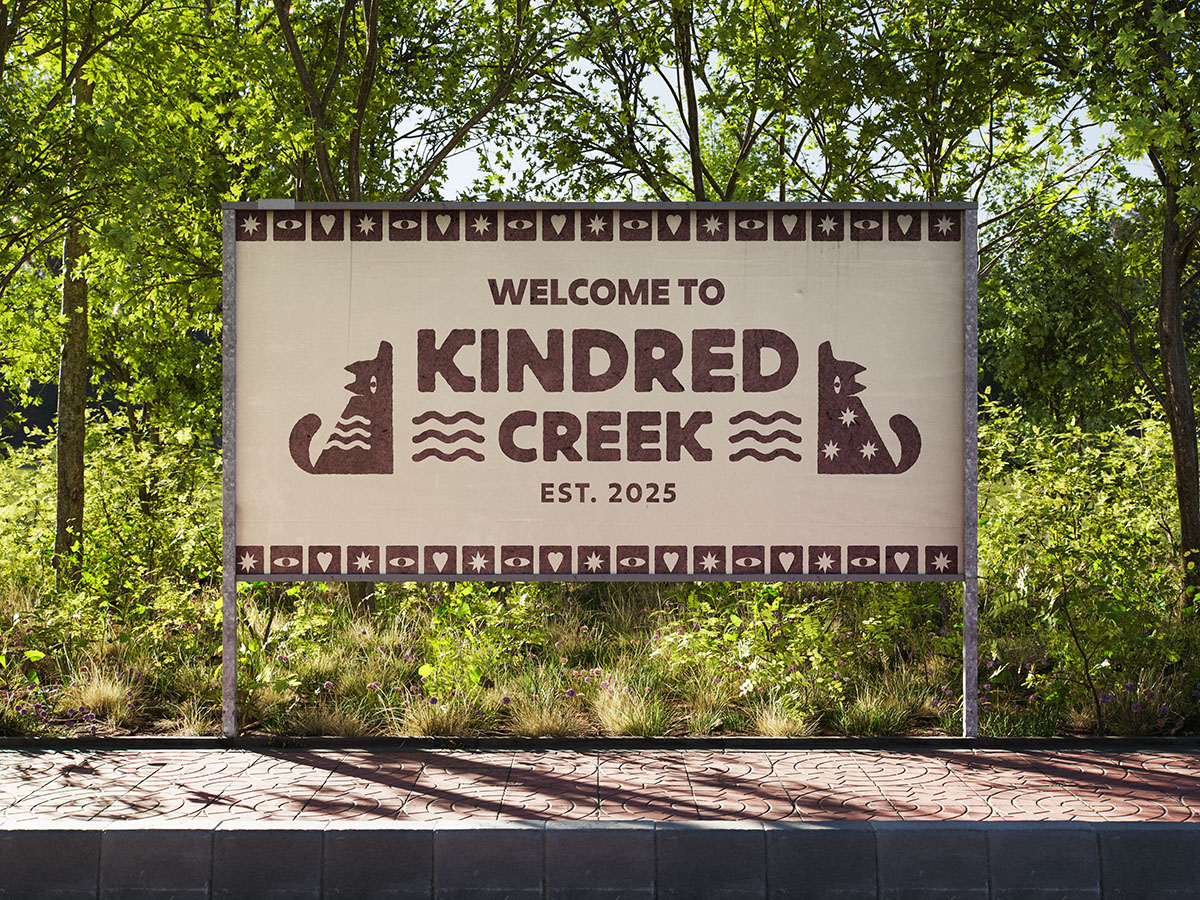 A large sign in anatural setting that says welcome to Kindred Creek. The billboard features simple, rustic illustrations of wolves, eyes, starts and water.
